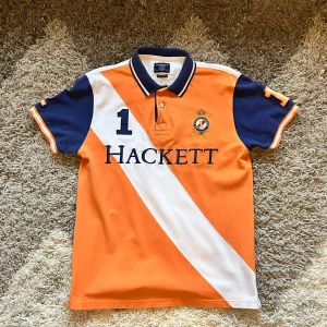 Orange pikétröja, Hackett - Vi säljer en snygg orange pikétröja från Hackett med vita och blå detaljer. Den har ett broderat emblem på bröstet och texten 'Beach Polo Team' på ryggen. Perfekt för en sportig look! Tröjan är i bomull och har korta ärmar.