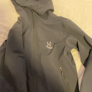 Svart vindjacka från Haglöfs - Säljer en svart vindjacka från Haglöfs i damstorlek M. Jackan har en praktisk huva och dragkedja framtill. Perfekt för blåsiga dagar och utomhusaktiviteter. Den är i mycket bra skick och redo för nya äventyr! Inga skador!