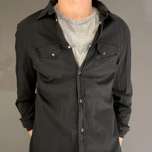 DONDUP SKJORTA - Säljer en stilren svart långärmad skjorta i bra skick. Skjortan har knappar framtill och två bröstfickor med knappdetaljer. Perfekt för både vardag och fest. Passar bra över en t-shirt för en avslappnad look, går även bra över skjorta. L passar M