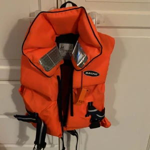 Orange flytväst från Baltic - Säljer en knallorange flytväst från Baltic, perfekt för barn mellan 15-30 kg. Västen har reflexdetaljer och justerbara remmar för säker passform. Den är i mycket bra skick och redo för nya äventyr på vattnet. 🧡