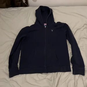GANT zip-up - Säljer en snygg mörkblå hoodie från GANT i storlek 176 cm, perfekt för tonåringar. Den har en dragkedja framtill och en klassisk huva. Tröjan är i mycket bra skick och passar perfekt för höst och vår. Superbekväm och stilren design!