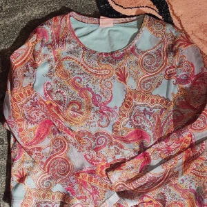 Mönstrad långärmad topp - Säljer en färgglad långärmad topp med ett livligt paisleymönster i rosa, orange och gult på en ljusblå bakgrund. Perfekt för att liva upp garderoben! Toppen har en lös passform och är gjord i ett mjukt material som känns skönt mot huden.