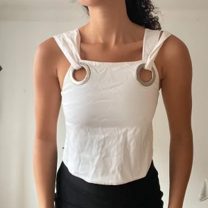 Vit ärmlös topp med ringdetaljer - Snygg vit ärmlös topp med unika ringdetaljer vid axlarna. Perfekt för sommardagar eller en utekväll. Toppen har en modern och stilren design som passar till både kjol och byxor. Materialet ser ut att vara bekvämt och lätt.