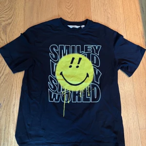 Svart smiley world T-shirt säljes - Svart Smiley world T-shirt säljes. På tröjan är det en gul smiley och det står smiley world. Tröjan är bara provad men aldrig använd. Storlek 170