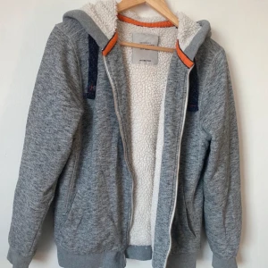 Grå fleecefodrad hoodie - Mysig grå hoodie från C&A med fleecefoder som håller dig varm. Den har en dragkedja framtill och en skön huva med snörning. Perfekt för kyliga höst- och vinterdagar. Storleken är inte angiven, men den ser ut att ha en normal passform.