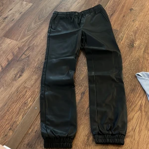 Svarta skinn byxor - Säljer ett par svarta skinn byxor med elastisk midja och muddar vid bensluten. Perfekta för en cool och avslappnad stil. Passar bra till både vardag och fest. 🖤