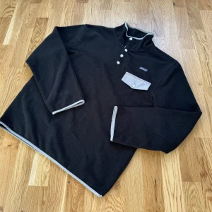 Svart fleece Synchilla från Patagonia - Säljer en svart fleece tröja från Patagonia i mycket bra skick. Den har en praktisk bröstficka med knapp och stilrena detaljer i grått. Perfekt för kyliga höst- och vinterdagar. Tröjan har långa ärmar och en bekväm passform. Passar både för vardag och friluftsliv.