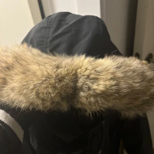 Svart parkajacka med pälsdetalj från Woolrich - Säljer en svart parkajacka från Woolrich i storlek XS. Jackan har en mysig pälsdetalj på huvan och stängs med både dragkedja och knappar. Perfekt för kalla vinterdagar! 🖤