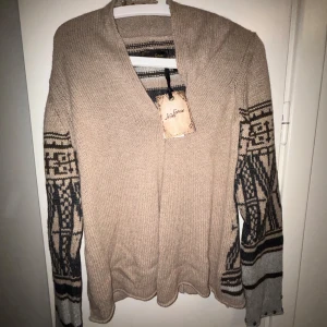 Beige stickad tröja från Non sense (oanvänd) - Säljer en superfin beige stickad tröja från Non sense . Den har ett unikt mönster i svart och grått på ärmarna och ryggen. Tröjan är långärmad och har en v-ringad design. Perfekt för höst och vinter, både mysig och stilren! 🌟