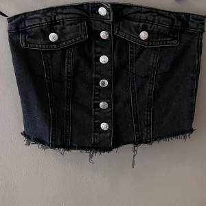 Svart jeanskorsett med knappar - Snygg svart jeanskorsett med knappar framtill och fransig nederkant. Den har en smockad detalj på baksidan för extra stretch och passform. Perfekt för en cool och trendig look!