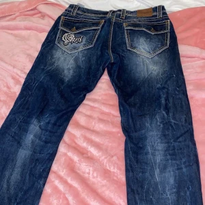 Blå jeans med broderad ficka - Säljer ett par snygga blå jeans med cool broderad detalj på bakfickan. De har en klassisk femficksdesign och slitna effekter för en trendig look. Perfekta för vardagsbruk eller en avslappnad utekväll. Passar bra med en t-shirt eller hoodie! Passar S/M. Tvättas innan utskick 