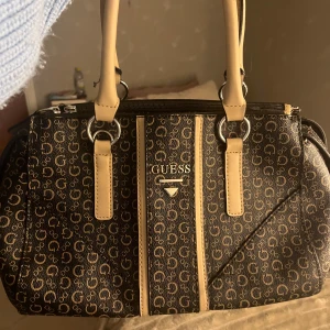 Vintage handväska från Guess - Säljer en snygg handväska från Guess som är vintage. Väskan har två  handtag och en dragkedja upptill. Insidan är rymlig med flera fack. Små defekter men inget som man tänker på tycker jag! (Pris kan diskuteras)
