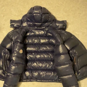 Mörkblå dunjacka från Moncler - Säljer en snygg och varm mörkblå dunjacka från Moncler. Jackan har en quiltad design och en praktisk huva. Perfekt för kalla vinterdagar. Den är i mycket bra skick och har en broderad Moncler-logga på ärmen. Insidan är brun och ger en fin kontrast NFC scan ingår.