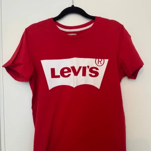 Röd Levi's t-shirt - Säljer en snygg röd t-shirt från Levi's i 100% bomull. Den har en klassisk passform med korta ärmar och det ikoniska Levi's-logotypen tryckt i vitt på framsidan. Perfekt för en casual look eller att bära till vardags. T-shirten är i mycket bra skick och redo för en ny ägare!
