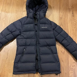 Mörkblå dunjacka från Peak Performance - Säljer en mörkblå dunjacka från Peak Performance i mycket bra skick. Jackan har en praktisk huva och dragkedja framtill. Perfekt för kalla vinterdagar och höstpromenader. Den har en stilren design med diskret logga på bröstet.