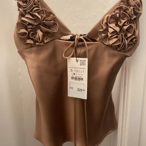 Beige satinlinne med volangdetaljer - Elegant beige satinlinne med justerbara smala axelband och vackra volangdetaljer vid bysten. Toppen har en knytning framtill och en mjuk, glansig finish som ger en lyxig känsla. Perfekt för festliga tillfällen eller en utekväll. Nytt med prislapp kvar!