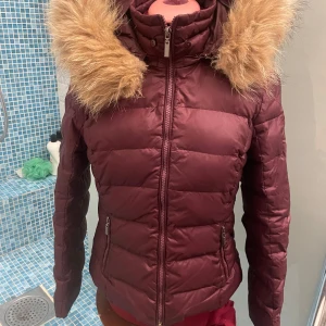 Vinröd dunjacka från Rockandblue - Säljer en snygg vinröd dunjacka från Rockandblue i storlek 38. Jackan har en fluffig fuskpäls på huvan och dragkedja framtill. Perfekt för kalla vinterdagar med sin varma och bekväma passform. Den är i mycket bra skick och redo för nya äventyr! ❄️har ett litet hål, där hyskan till en av knappar till pälskanten släppt, lätt att sy ihop