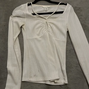 Vit ribbad långärmad topp från H&M - Säljer en stilren vit ribbad topp från H&M. Den har långa ärmar och en snygg v-ringad design med knytning framtill. Perfekt för både vardag och festliga tillfällen. Passar bra till jeans eller kjol för en chic look!