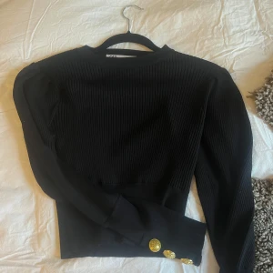 Svart ribbad tröja från Zara - Snygg svart ribbad tröja från Zara med långa ärmar och guldknappar vid ärmsluten. Perfekt för både vardag och fest. Tröjan har en elegant och stilren design som passar till det mesta. 🖤 