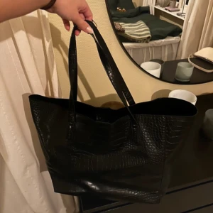 Svart axelväska i syntet - Säljer en snygg svart axelväska i syntetmaterial med krokodilmönster. Perfekt för både vardag och fest! Väskan har en stilren design och är rymlig nog för alla dina nödvändigheter. Den är i mycket bra skick och redo för en ny ägare. Den har lite skador (se andra bilden)