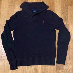 Svart stickad tröja från Ralph Lauren - Säljer en snygg svart/mörkblå stickad tröja från Ralph Lauren med en röd logga på bröstet. Tröjan har en unik krage med knappar och långa ärmar. Perfekt för kyliga dagar och ger en stilren look. Passar både till vardags och lite finare tillfällen. 