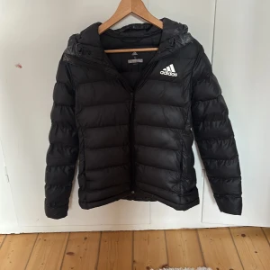 Svart dunjacka från Adidas - Säljer en svart dunjacka från Adidas i mycket bra skick (endast några smink fläckar som går bort i tvätten).Jackan har en dragkedja framtill och en justerbar huva. Perfekt för kyliga dagar och håller dig varm och bekväm. Passar både till vardags och sportiga aktiviteter.