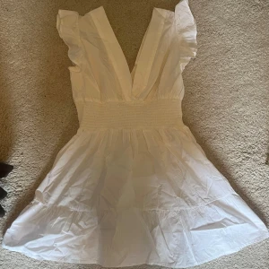 Vit kortklänning med volangdetaljer - Säljer en superfin oanvänd vit kortklänning med volangdetaljer och djup v-ringning. Den har en smockad midja som ger en smickrande passform. Klänningen är ärmlös och gjord i ett lätt och luftigt material.