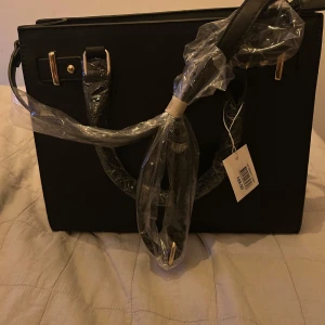 Svart handväska med gulddetaljer - Elegant svart handväska med gulddetaljer och dragkedja. Perfekt för en stilren look. Väskan har både handtag och axelrem för flexibilitet.