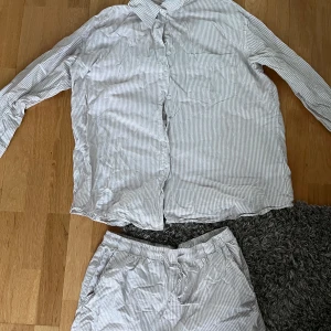 Randig pyjamasskjorta och shorts från Lager 157 - Säljer en bekväm randig pyjamasskjorta med matchande shorts från Lager 157. Setet är i ljusgrått och vitt med knappar på skjortan och elastisk midja på shortsen. Perfekt för en avslappnad dag hemma.