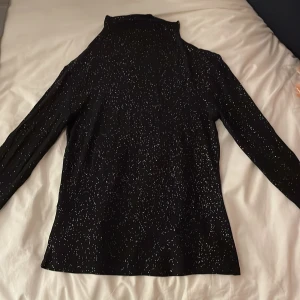 Svart glittrig polotröja från Vila - Säljer en svart polotröja från Vila med glittrigt mönster. Tröjan har långa ärmar och en elegant polokrage, perfekt för att lysa upp din garderob.