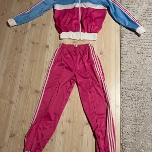 Tracksuit original vintage från Adidas - Säljer en retro original tracksuit från Adidas i blått och rosa med vita detaljer. Byxor med mudd å en ficka .Jackan har dragkedja,fickor och långa ärmar, perfekt för en sportig look. Den är tillverkad i polyester och har det klassiska Adidas-märket broderat på bröstet. Vintage i nyskick