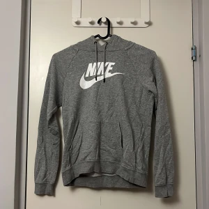 Grå hoodie från Nike - Säljer en grå hoodie från Nike med klassisk logga i vitt på bröstet. Tröjan har en bekväm passform med långa ärmar och en praktisk huva med snören. Perfekt för en avslappnad stil.