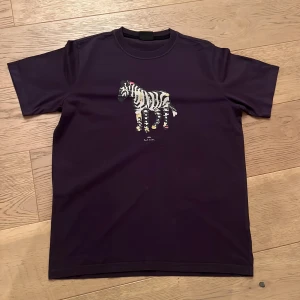 Lila  t-shirt med zebra från Paul Smith - Snygg lila  t-shirt från Paul Smith med ett coolt zebramotiv på framsidan. Perfekt för en avslappnad stil med en touch av unik design. T-shirten har korta ärmar och är gjord i mjukt material.