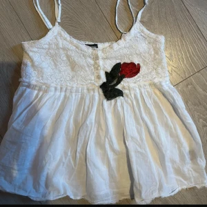Linne - Söt vit topp  med spetsdetaljer och en broderad röd ros framtill. Toppen har knappar framtill och tunna axelband, perfekt för varma dagar. Tröjan sitter tajt vid bröstet och lite fladdrig nedtill. Står att den är storlek M, men den passar XS-S och mindre. Använd 1 gång🥀