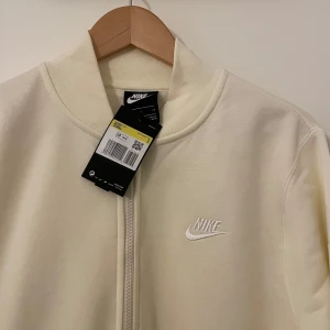 Gul/vit kofta från Nike - Säljer en helt oanvänd stilren kofta från Nike med dragkedja och broderad logga på bröstet. Plagget har långa ärmar, praktiska fickor och en insida gjort av fleecematerial. Perfekt för vardagsbruk.