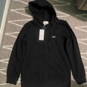 Svart hoodie från Lacoste - Säljer en stilren svart hoodie från Lacoste med dragkedja och den klassiska krokodilloggan på bröstet. Perfekt för en avslappnad look. Hoodien har långa ärmar och en praktisk huva med dragsko.