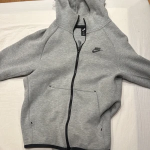 Nike tech - Säljer en grå huvtröja från Nike med dragkedja och huva. Tröjan har långa ärmar och två fickor framtill. Perfekt för en avslappnad stil.