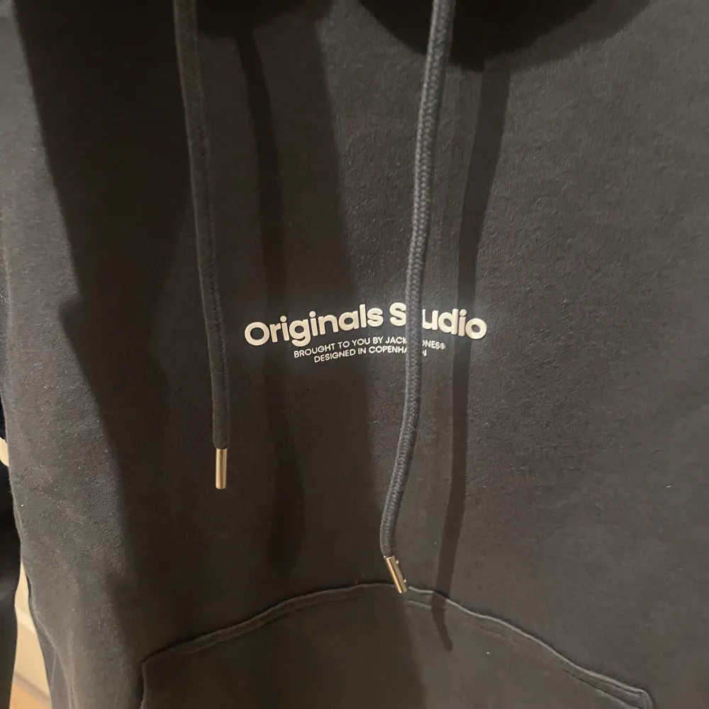 Säljer en snygg mörkgrå hoodie från Jack & Jones med texten 'Originals Studio' på bröstet. Den har en klassisk känguruficka och justerbar dragsko i huvan. Perfekt för en avslappnad stil.. Neuletakit & Villapaidat.