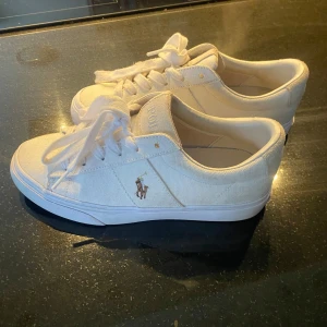 Vita sneakers från Polo Ralph Lauren - Snygga vita sneakers från Polo Ralph Lauren med klassisk design och broderad logotyp på sidan. Skorna har snörning och är tillverkade i textilmaterial, vilket ger en stilren och bekväm känsla.