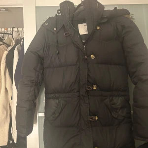 Svart dunjacka från Boomerang - Säljer en svart dunjacka från Boomerang i storlek XXS. Jackan har en klassisk design med knappar och dragkedja, samt praktiska fickor. Perfekt för kalla dagar med sin varma dunstoppning.