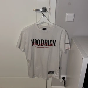 Vit t-shirt från Hoodrich - Snygg vit t-shirt från Hoodrich med svart och röd logga på bröstet. Den har korta ärmar och en cool detalj med en etikett längst ner. Perfekt för streetwear-stilen! Pris kan diskuteras.