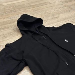 Svart hoodie från Polo Ralph Lauren - Snygg svart hoodie från Polo Ralph Lauren med dragkedja och klassisk logga på bröstet. Tröjan har en justerbar huva och praktiska fickor framtill. Perfekt för en avslappnad stil. Endast: 550