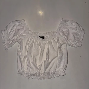 Vit offshoulder blus - Säljer en söt vit offshoulder blus från Gina tricot. Blusen har puffiga korta ärmar och ett elastiskt band runt hals och midja för en bekväm passform. Den är dekorerad med små broderade detaljer som ger en romantisk känsla.