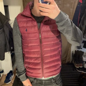 Vinröd dunväst från Polo Ralph Lauren - Snygg vinröd dunväst från Polo Ralph Lauren med dragkedja och broderad logga på bröstet. Perfekt för lager-på-lager under höst och vår. Västen har en quiltad design och praktiska fickor med dragkedja. Cond 10/10