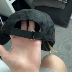 Svart keps med mönster och röd-gröna detaljer - Snygg svart keps med diskret mönster och röd-gröna ränder på sidan. Justerbar rem baktill för perfekt passform. Perfekt för en stilren look.