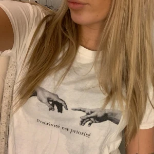 Vit t-shirt med tryck - Vit t-shirt med ett konstnärligt tryck av två händer och texten 'Positivité est priorité' på framsidan. 