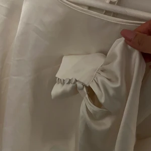 Vit satinblus med knytning - Elegant vit blus i satin med långa ärmar och knytning i ryggen. Blusen har en mjuk och glansig yta som ger en lyxig känsla. Perfekt för en stilren look.