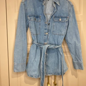 Jeansjacka med bälte från Bubbleroom - Snygg jeansjacka från Bubbleroom i ljusblå denim. Jackan har långa ärmar, knappar framtill och två bröstfickor med lock. Den är utrustad med ett knytbälte i midjan för en figurnära passform.