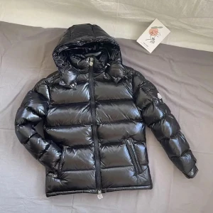 Moncler maya  - Snygg svart pufferjacka från Moncler med glansig finish och dragkedja framtill. Jackan har en praktisk huva och två fickor med dragkedjor. Perfekt för kyliga dagar.