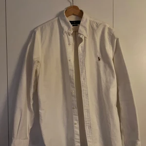 Ralph lauren skjorta - Säljer en klassisk vit skjorta från Ralph Lauren i slim fit. Skjortan har långa ärmar och knappar framtill samt en broderad logotyp på bröstet. Perfekt för en stilren look. Väldigt lite använd och ser helt ny ut! Tvättad endast 1 gång. Hör av er vid frågor! OBS! Vid en snabb affär är priset inte hugget i sten!😊
